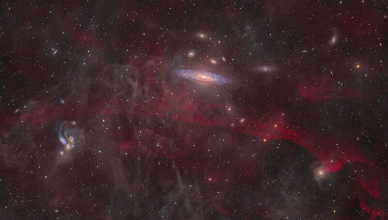 A deep space image showing several galaxies among countless stars, with wispy red nebulae stretching across the scene and faint clouds of interstellar dust in the background.