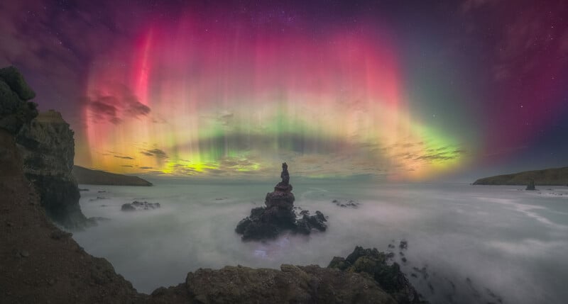 A vibrant aurora borealis glows in pink, green, and yellow hues over a rocky seashore at night, with misty waves and a rock formation at the center under a starry sky.