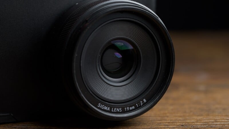 Close-up of a Sigma 19mm f/2.8 camera lens attached to a black camera, with the lens glass and textured focus ring visible, set against a wooden surface.