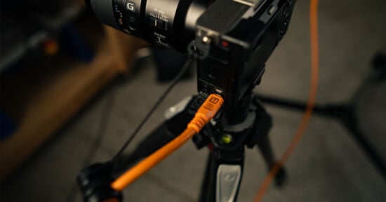 A close-up of a camera mounted on a tripod with an orange USB cable plugged into its side, likely for tethered shooting; the background is blurred.