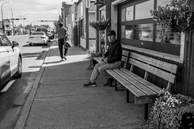 A man wearing a hat sits alone on a wooden bench outside a building on a city sidewalk while another person walks away in the background; parked cars and plants are also visible. The image is in black and white.