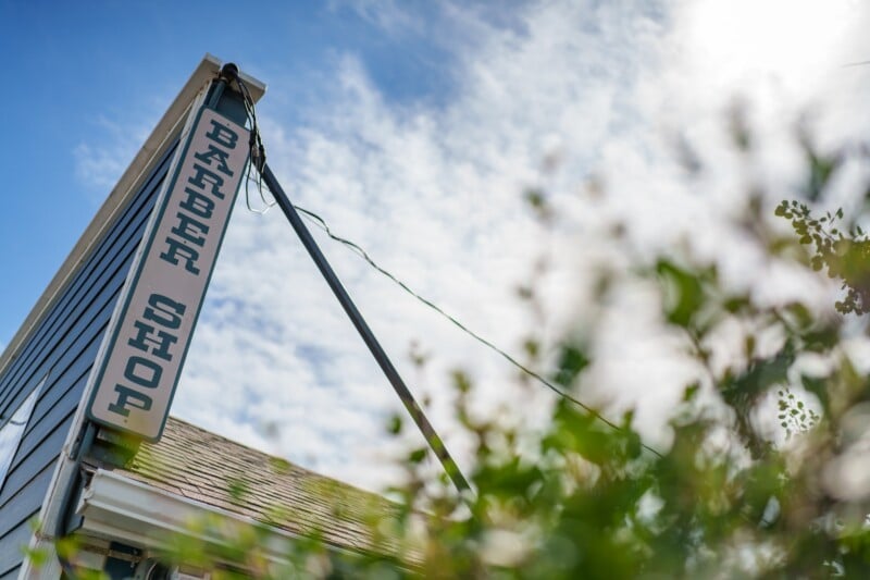 A close-up of a vintage "Barber Shop" sign attached to the corner of a building, with green foliage in the foreground and a bright, partly cloudy sky in the background.
