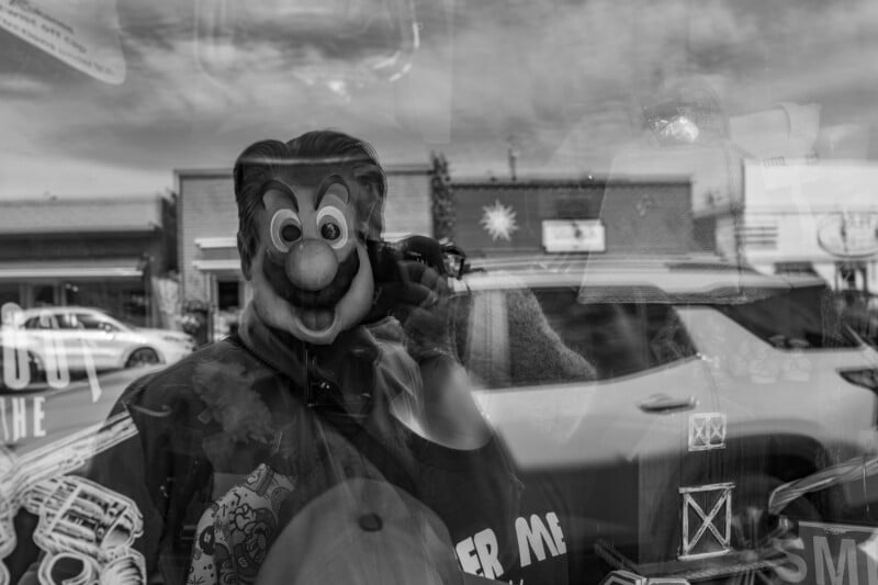 A person wearing a cartoonish mask is seen through a shop window, with reflections of cars, buildings, and street elements blending into the scene. The photo is in black and white, creating a surreal, layered effect.