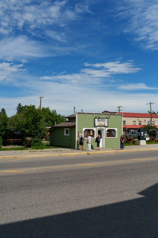 Four people stand outside a small green building called "Prairie Donair" on a sunny day, with trees and other buildings in the background and a clear blue sky overhead.