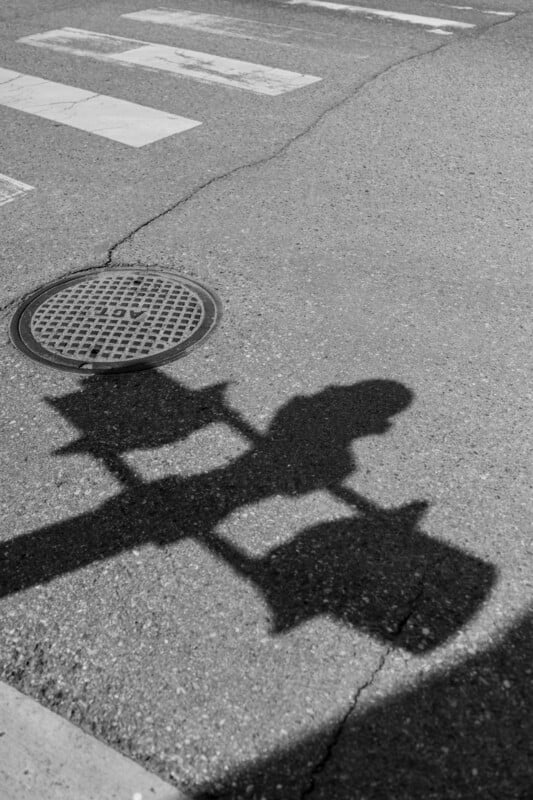 Black and white photo of a street with a crosswalk, a manhole cover, and the shadow of a traffic light cast on the pavement.