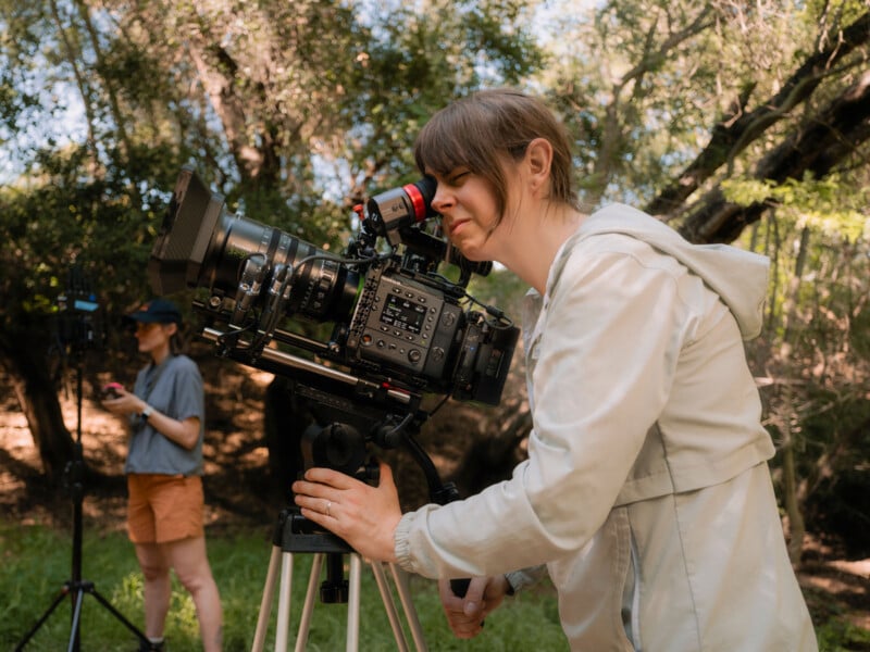 A person in a light jacket looks through a professional video camera on a tripod outdoors, while another person stands in the background with camera equipment among trees.