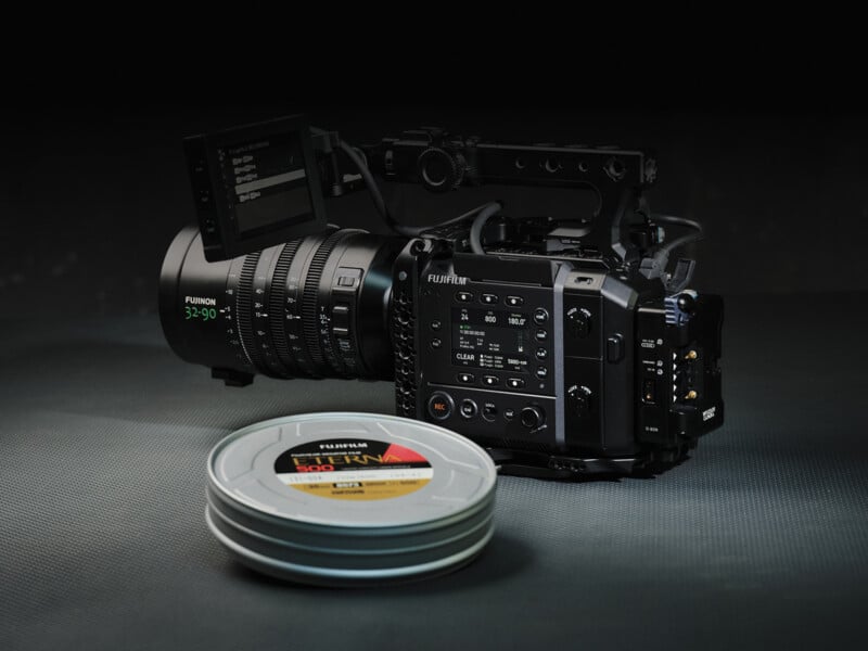 A professional FUJIFILM cinema camera with a large lens and side monitor sits on a textured surface, next to two stacked metal film canisters labeled “ETERNA.”.