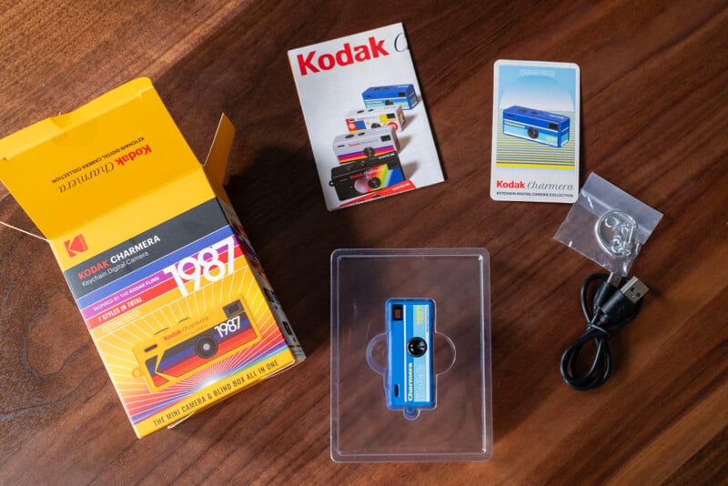 Kodak-themed items on a wooden table: an open yellow box, a blue mini camera, two instruction booklets, a USB cable, a keychain, and clear plastic packaging.