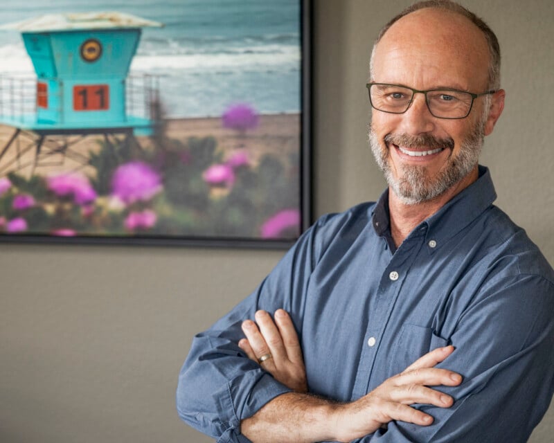 A smiling man with glasses and a beard stands with arms crossed in front of a wall featuring a framed photograph of a beach scene and a turquoise lifeguard tower.