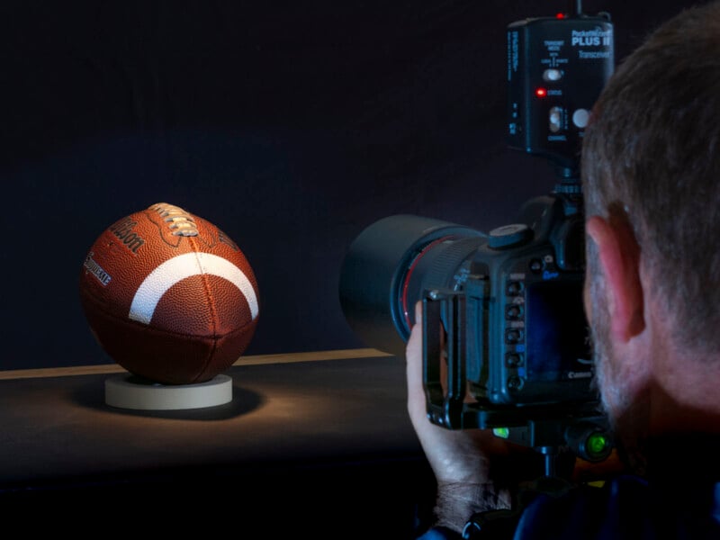 A photographer takes a close-up photo of an American football placed on a small stand, using a professional camera with a large lens against a dark background.
