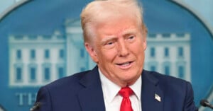 A man in a navy suit, white shirt, and red tie speaks at a podium, with a blurred image of the White House in the background.