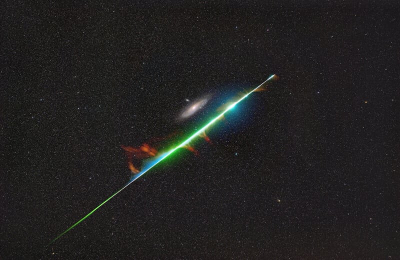 A bright green meteor streaks diagonally across a star-filled night sky, with a galaxy visible in the background; the meteor glows and leaves a vivid colorful trail.