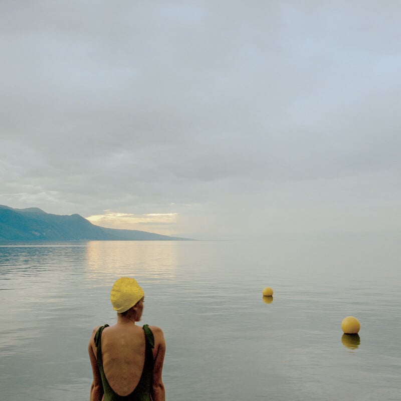 A person in a swimsuit and yellow swim cap stands at the edge of a calm, expansive lake with three yellow buoys floating in the water, mountains in the distance, and a cloudy sky overhead.