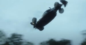 A black car is airborne, flipping high above the ground against a cloudy sky, with trees blurred below, suggesting fast motion and an intense action scene.
