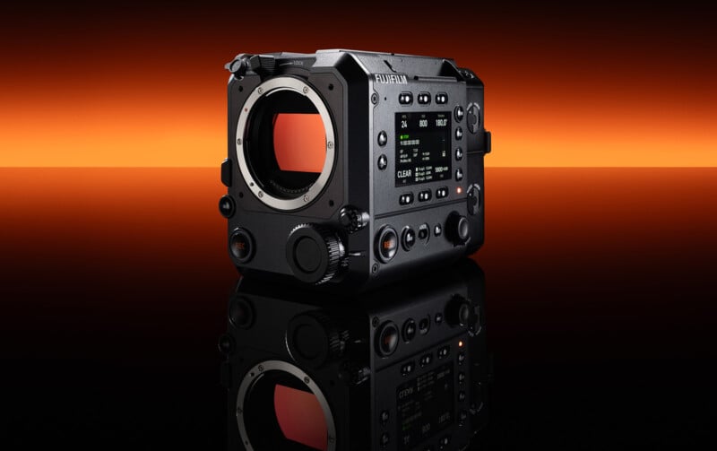 A black Fujifilm cinema camera body with a lens mount and digital display is shown on a reflective surface. The background features a gradient from deep orange to black.