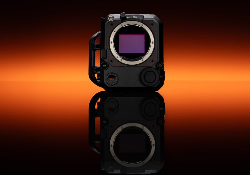 A professional digital cinema camera body with exposed sensor is displayed upright on a reflective surface against a vibrant orange gradient background.