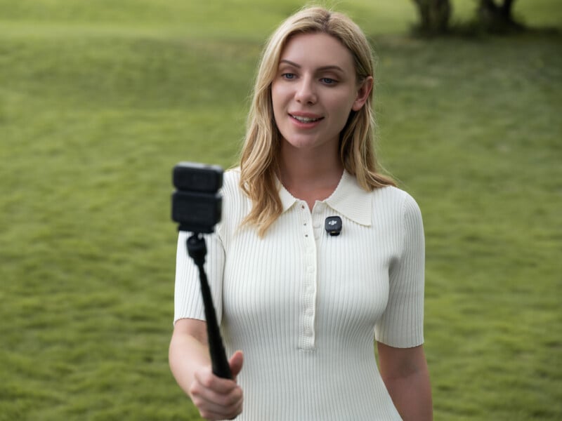 A woman wearing a light-colored dress stands outdoors on grass, holding a selfie stick with a camera attached, smiling and looking at the device.