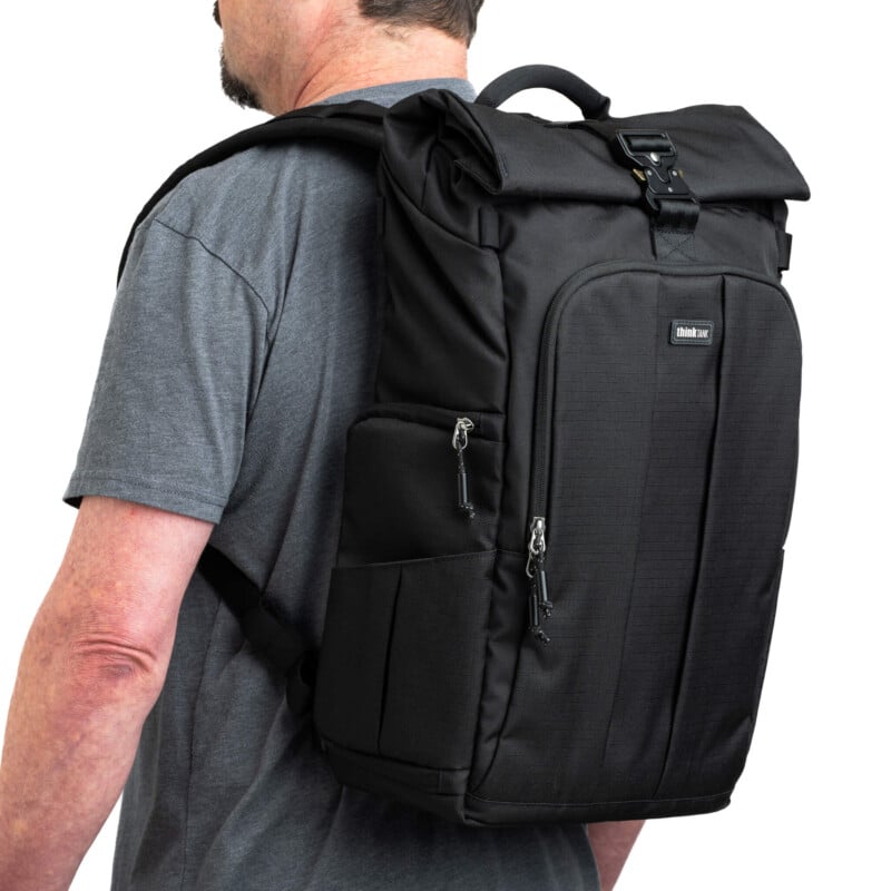 Un hombre con una camiseta gris con una mochila negra y negra rodante con múltiples compartimentos con cremallera y correas de hombro acolchadas.