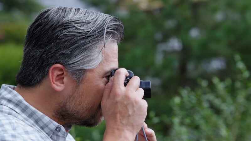 A man with salt-and-pepper hair and a beard holds a camera up to his eye, taking a photo outdoors. He is wearing a plaid shirt, and green foliage is blurred in the background.