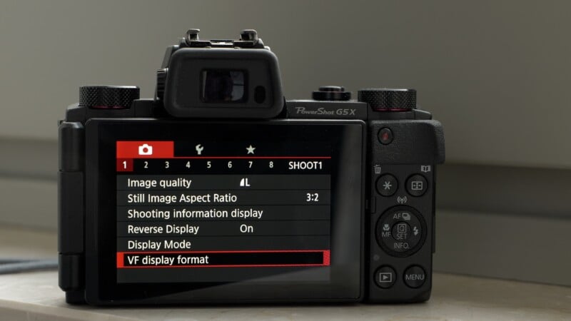 A digital camera sits on a surface with its screen displaying the shooting settings menu, highlighting "VF display format" in red, alongside options for image quality, aspect ratio, and display settings.