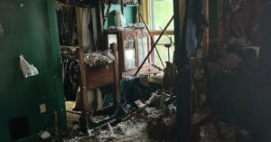 A room with green walls is heavily damaged, with debris and insulation scattered on the floor and furniture. A broken window lets in light, and a wooden cabinet and various household items are covered in rubble.