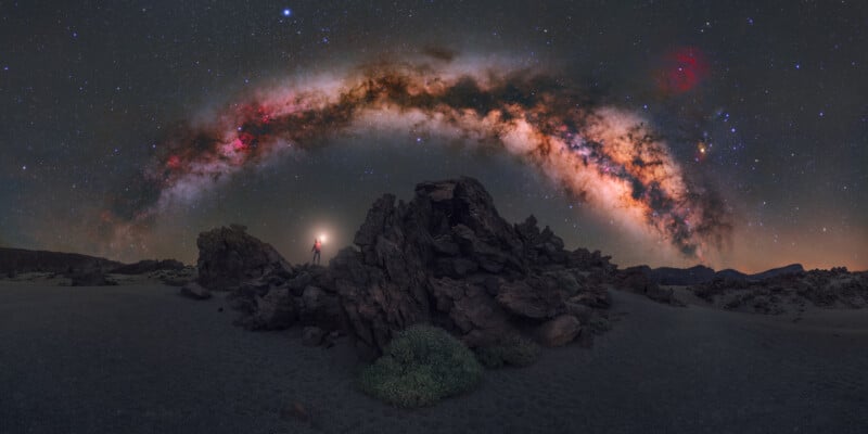 A person stands on rugged rocks beneath a vibrant Milky Way arch stretching across a starry night sky in a remote, rocky desert landscape.