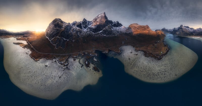Aerial view of a rugged, mountainous coastline with snow-dusted peaks, a winding road, and shallow turquoise waters meeting deep blue sea under a dramatic, cloudy sky at sunrise or sunset.