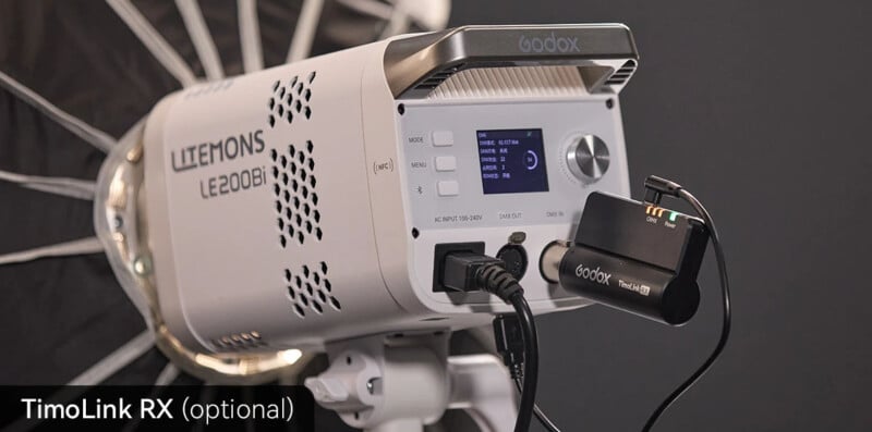Close-up of a Godox Litemons LE200Bi studio light with control panel, display screen, and connected TimoLink RX wireless receiver in a photography setup. A large reflective umbrella is visible in the background.