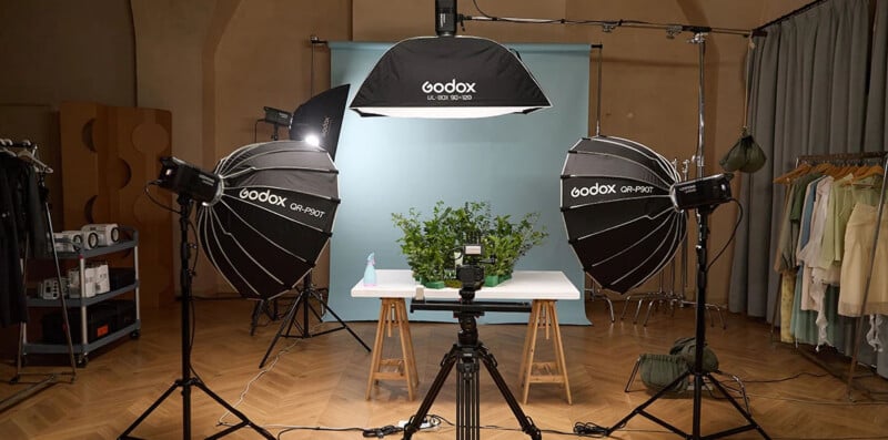 A professional photography setup with three Godox studio lights aimed at a table with green plants, a camera on a tripod, and a blue backdrop. Clothing racks and equipment are visible in the background.