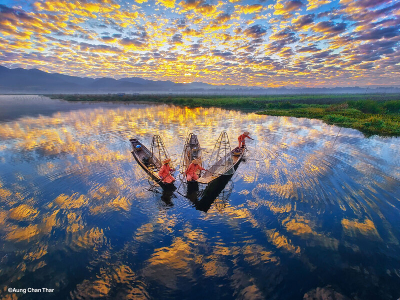 Tres pescadores en un bote tradicional proyectan una red en un lago reflectante al amanecer, con vibrantes nubes y colores en el agua, rodeados de vegetación y montañas distantes.