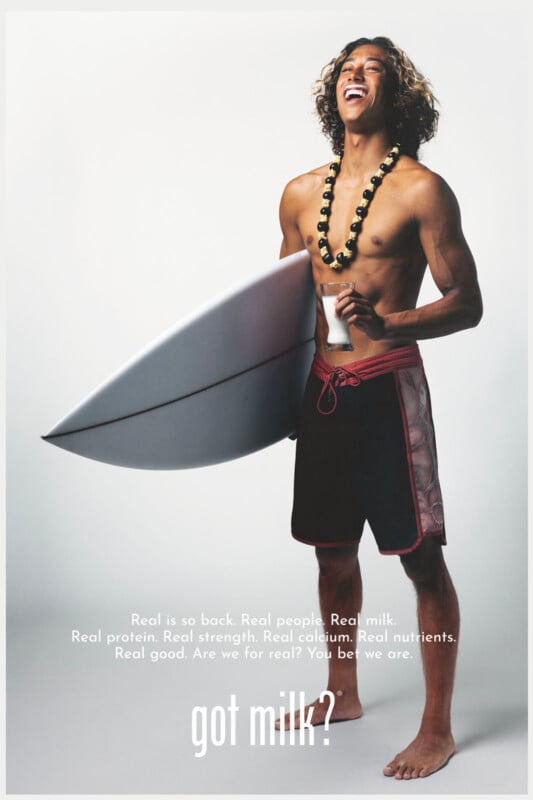 Un joven sonriente sosteniendo una tabla de surf y una botella de leche con puños desnudos, con pantalones cortos de madera y collar con cuentas. El texto en la parte inferior dice: 