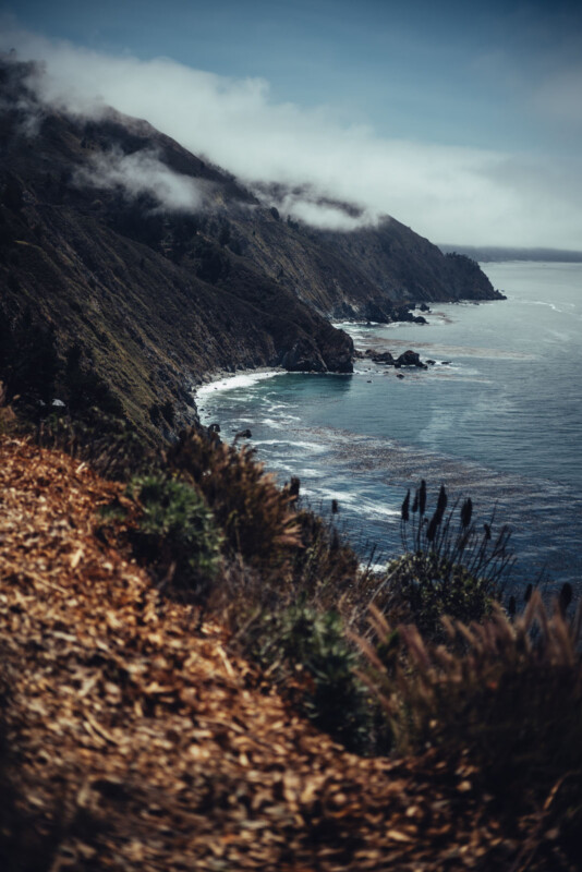 La robusta costa, los empinados acantilados cubiertos de vegetación, encontrando el océano azul debajo. Las olas blancas se estrellaron a lo largo de la orilla. Las nubes bajas en la cima de la montaña están en la cima de la montaña, y algunas plantas costeras son visibles en primer plano.
