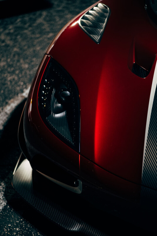 Primer plano de los faros izquierdos y la capucha de un elegante automóvil deportivo rojo con detalles de fibra de carbono que reflejan la luz en un entorno tenue.