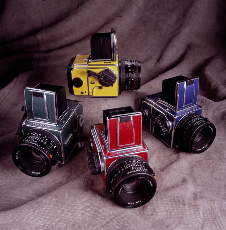 Four Hasselblad cameras with colorful bodies—green, red, blue, and yellow—are arranged on a draped, gray fabric background. Each camera features a waist-level viewfinder and classic medium-format design.