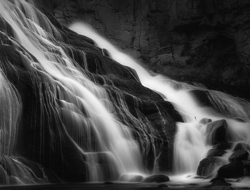 A black and white photo of a cascading waterfall flowing over dark, jagged rocks, with soft, misty water creating a smooth, silky effect. The background shows rocky cliffs and sparse vegetation.