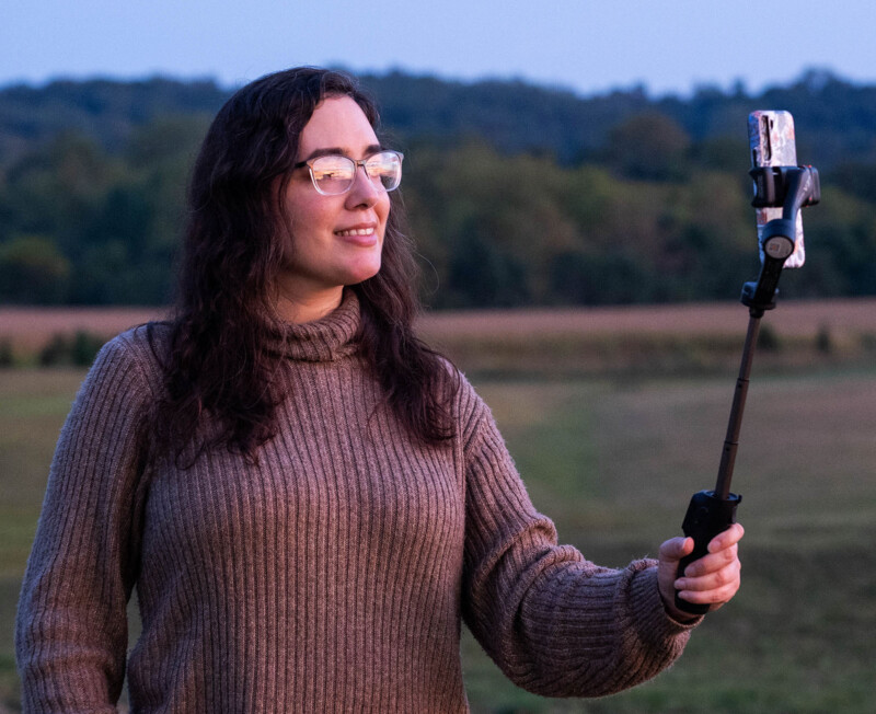 A woman with long brown hair and glasses, wearing a brown sweater, smiles while holding a smartphone on a selfie stick outdoors with fields and trees in the background.