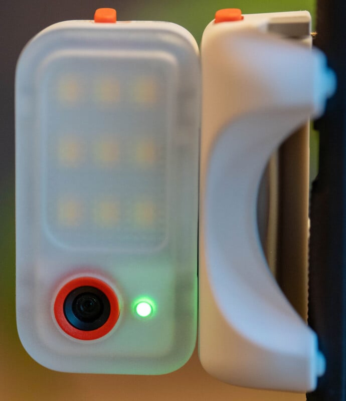 Close-up of a small device with a camera lens, LED lights, and a glowing green indicator, attached to a white plastic clip with orange and blue buttons.