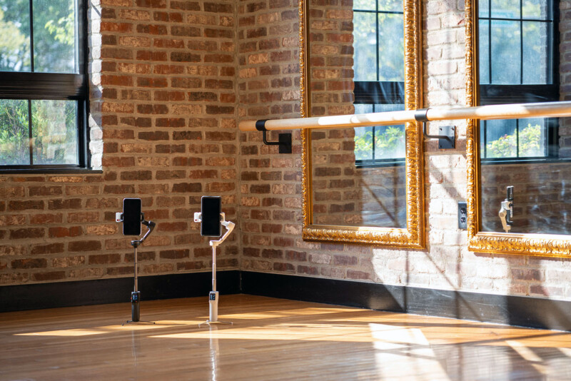 A bright dance studio with exposed brick walls, large mirrors with gold frames, a ballet barre, hardwood floors, and two phone mounts positioned on stands near the mirrors. Sunlight streams through the windows.