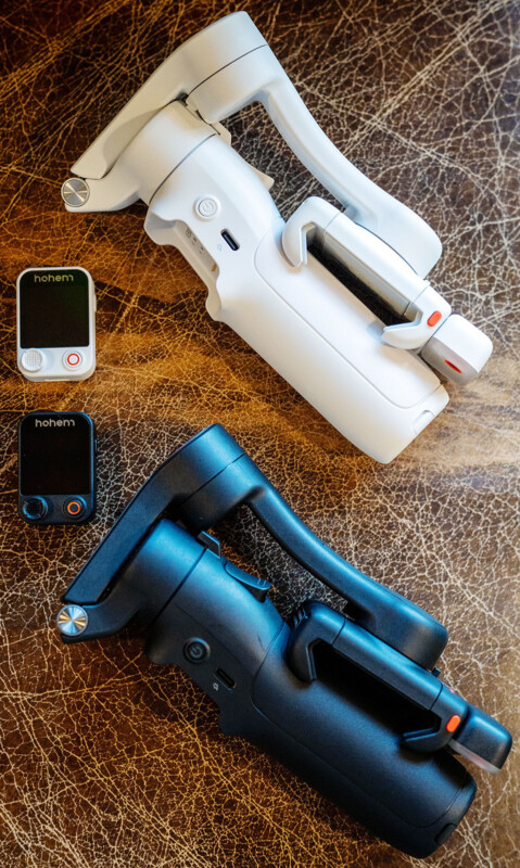 Two handheld gimbals, one white and one black, are placed on a brown textured surface. Next to each gimbal is a matching remote control labeled "hohem," one in white and one in black.