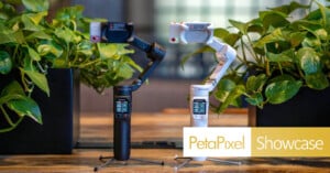 Two handheld gimbals, one black and one white, are standing on a wooden table surrounded by green plants. Both have small screens and are branded "Hohem." "PetaPixel Showcase" text is displayed in the corner.