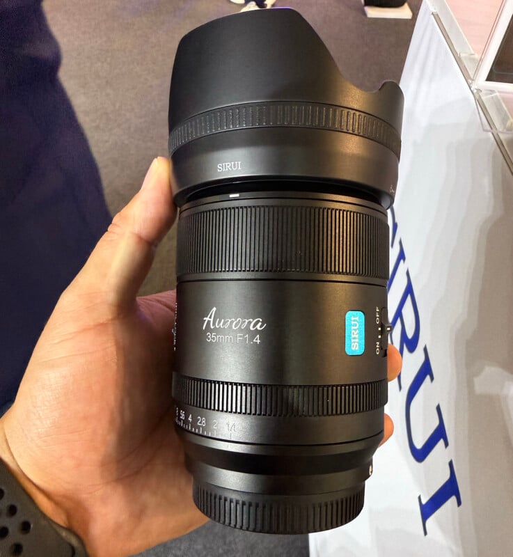 A hand holding a Sirui Aurora 35mm F1.4 camera lens displayed vertically, with a petal-shaped lens hood attached and branding clearly visible on the lens barrel.