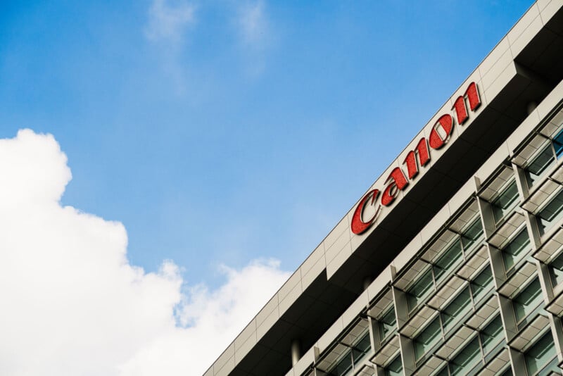 A modern office building with the Canon logo in red on the upper corner, set against a blue sky with scattered white clouds.