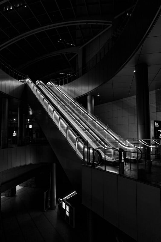 A black and white image of an empty, illuminated escalator in a modern indoor space, surrounded by curved railings and architectural details. The scene is devoid of people and has a dramatic, moody atmosphere.
