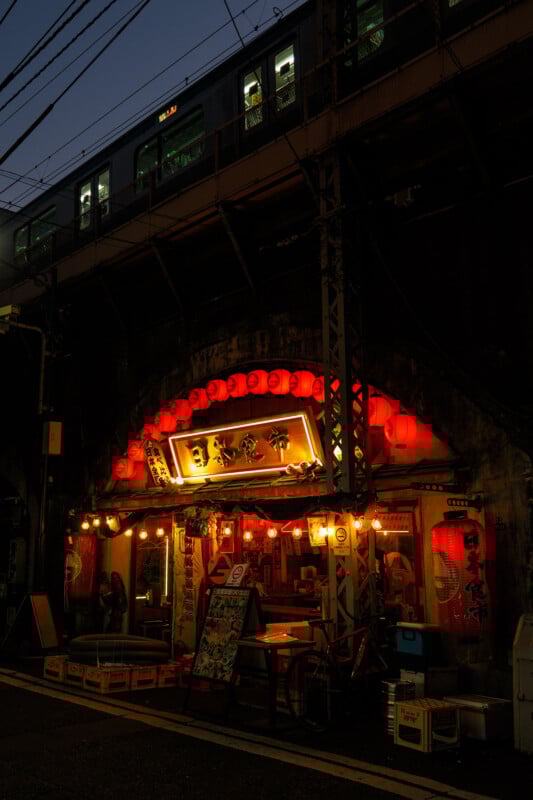 A small Japanese restaurant with red lanterns glowing under a railway bridge at dusk; a train passes overhead, while outdoor tables, signs, and a bicycle are visible in front of the entrance.