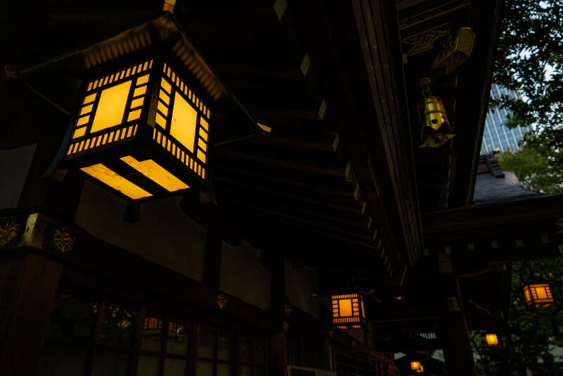 Traditional Japanese lanterns glow warmly under the eaves of a wooden building, with intricate architectural details visible. Trees and a modern high-rise are faintly seen in the background.