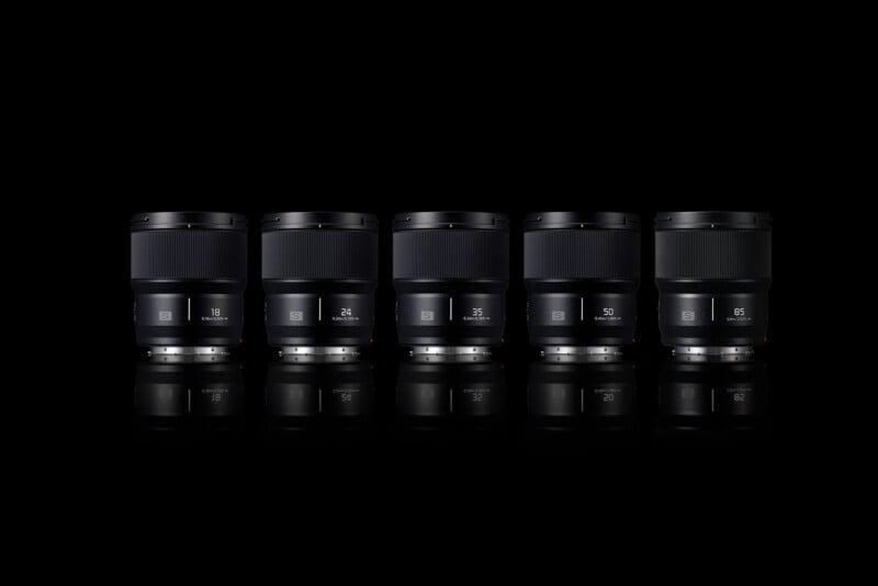 Five camera lenses are arranged side by side against a black background, each displaying different focal lengths. The lenses and their reflections create a symmetrical, minimalist composition.