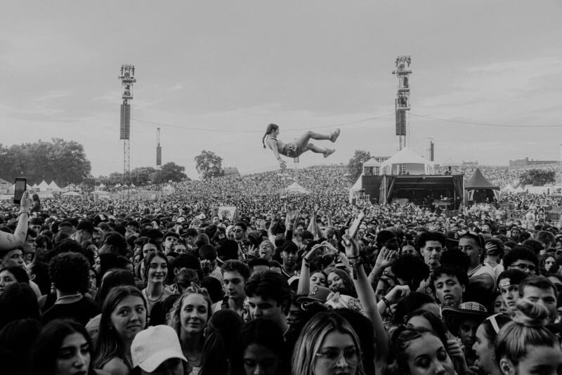 En un festival de música al aire libre, hay una multitud de personas navegando por la audiencia. Esta foto es en blanco y negro, con el escenario y la torre visibles en el fondo.