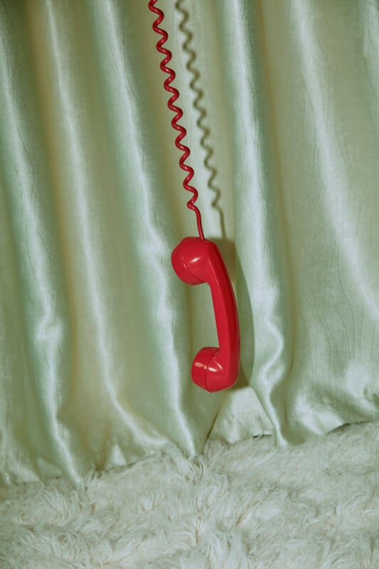 A red corded telephone receiver hangs in front of shiny, light-colored curtains and a fluffy white carpet.