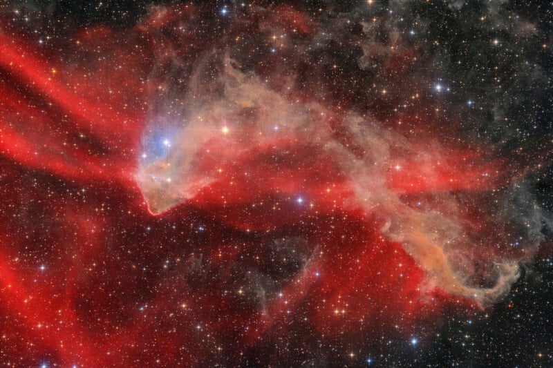 Brilliant red and orange nebula clouds stretch across a star-filled background in deep space, with wispy, ethereal patterns illuminated by bright stars.