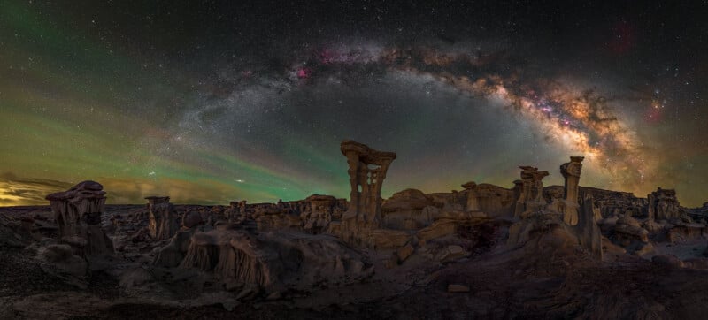 Rocky hoodoo formations rise under a vivid night sky, with the Milky Way arching overhead and colorful aurora-like lights illuminating the landscape in a remote desert scene.